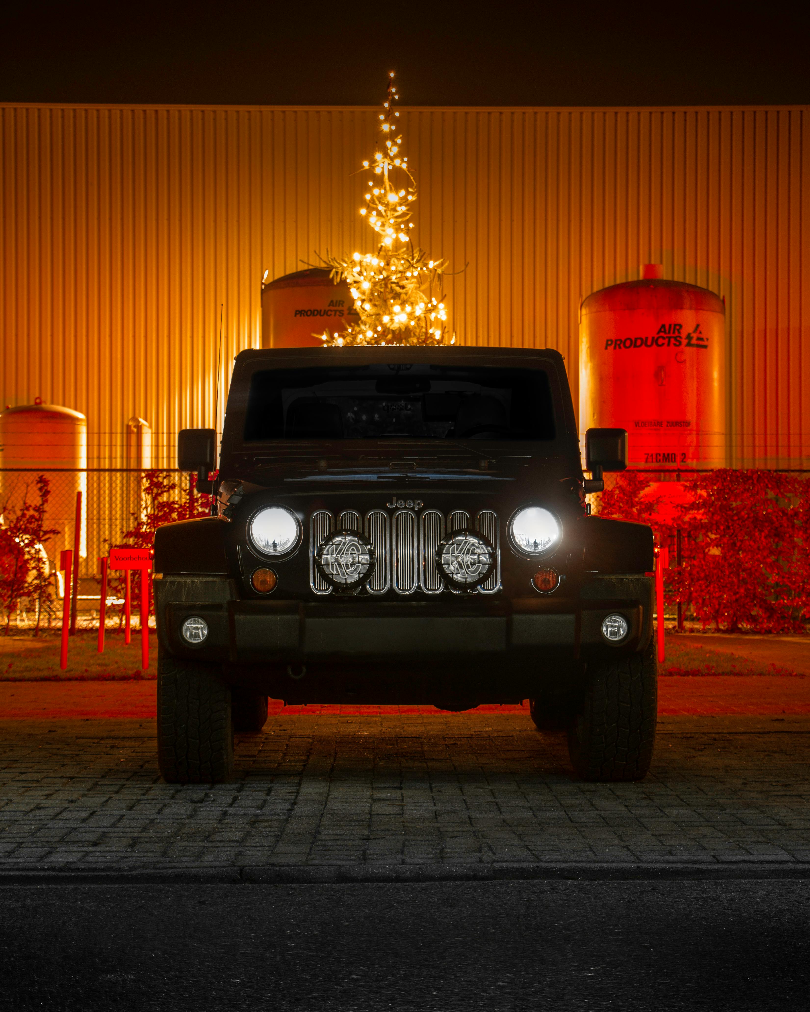 A Jeep parked outdoors at night, highlighted by festive lighting and industrial backdrop.
