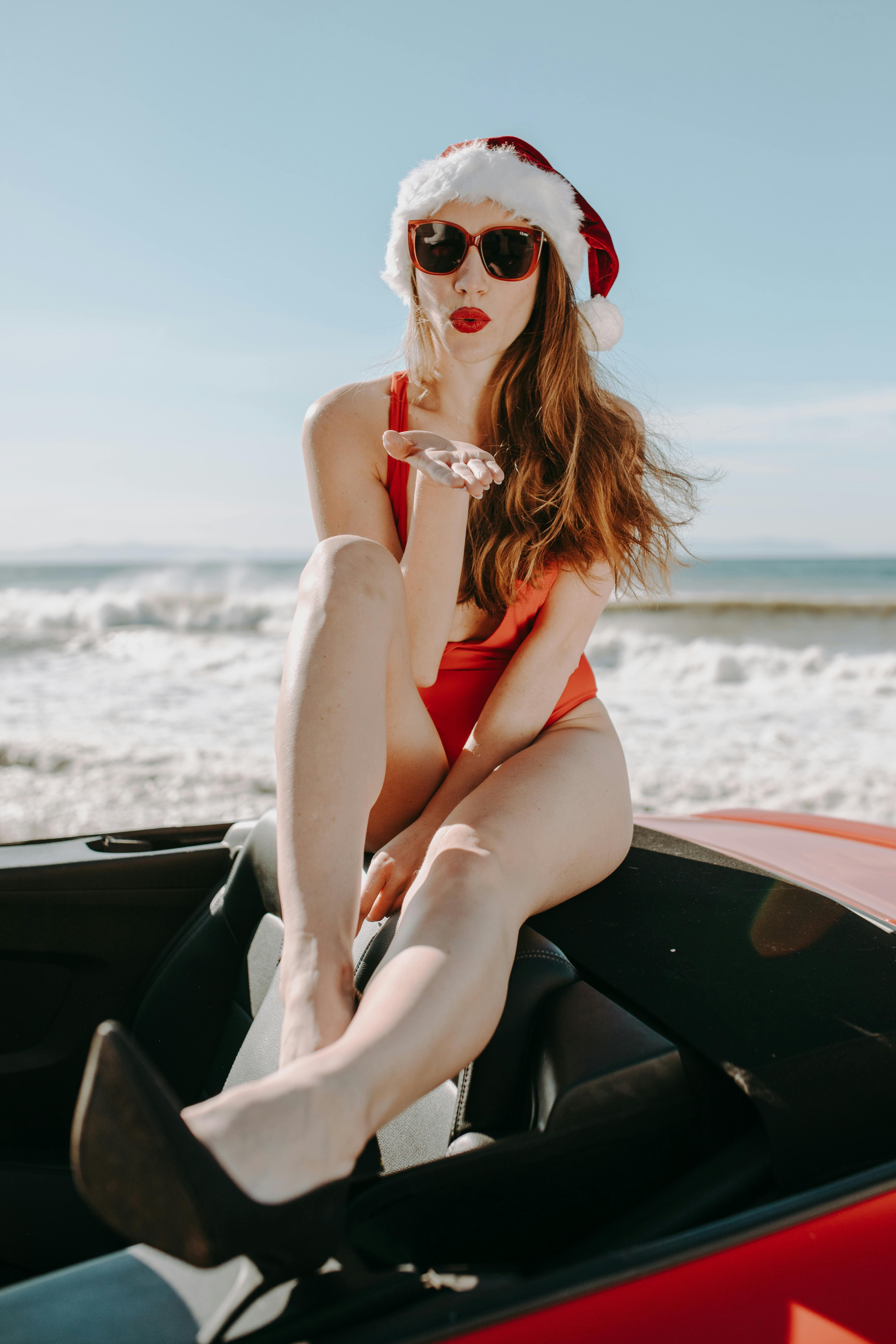 Woman in Santa hat and swimwear posing on a beach with a convertible.